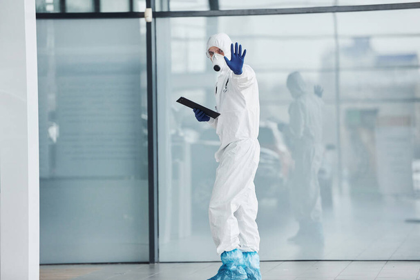 Shows stop gesture by hand. Male doctor scientist in lab coat, defensive eyewear and mask holding notepad in hands. - 写真・画像