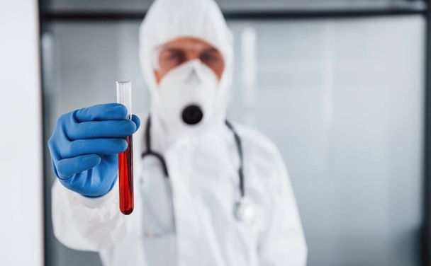 Male doctor scientist in lab coat, defensive eyewear and mask holds test tube with blood. - 写真・画像
