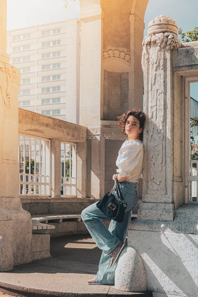 Portrait of carefree teenage girl posing for camera, squinting eyes from rays of sun, standing in relaxed pose near column of marble gazebo in park, wearing loose-fitting denim trousers and an elegant blouse - Foto, afbeelding