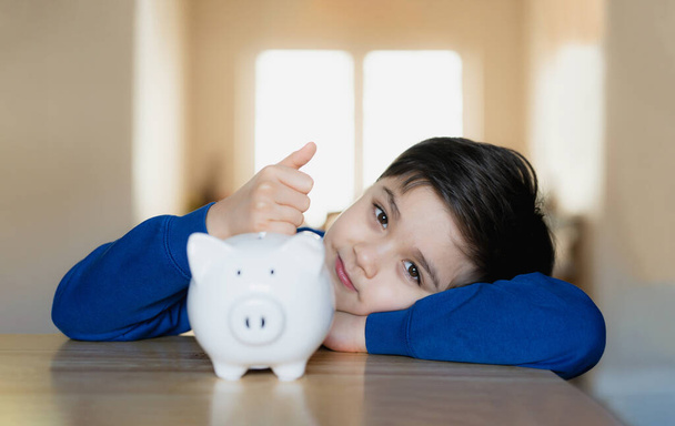 Happy School boy putting thumbs up on piggy bank and looking at camera with smiling face.Child showing money saving box.kid Learning financial responsibility and planning about saving money for future - Photo, Image