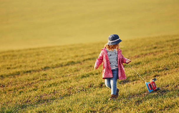 Cute little girl walks with toy car on the beautiful field at sunny daytime. - Photo, Image