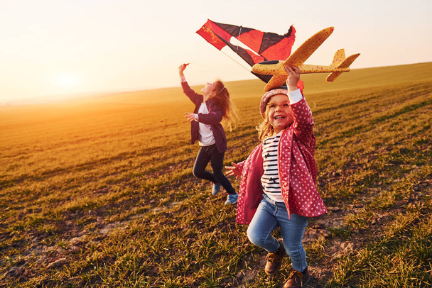Two little girls friends have fun together with kite and toy plane on the field at sunny daytime. - Fotoğraf, Görsel