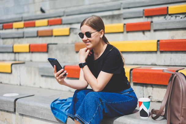 Портрет расслабленной молодой женщины, сидящей снаружи в городе со своим смартфоном, смотрящей в сторону и улыбающейся - Фото, изображение