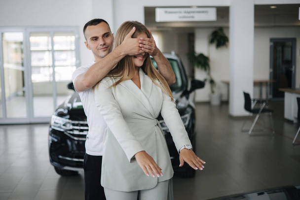 Man close eyes to his wife and makes a surprise buying car. Man and woman in car showroom. Happy young couple - Photo, image
