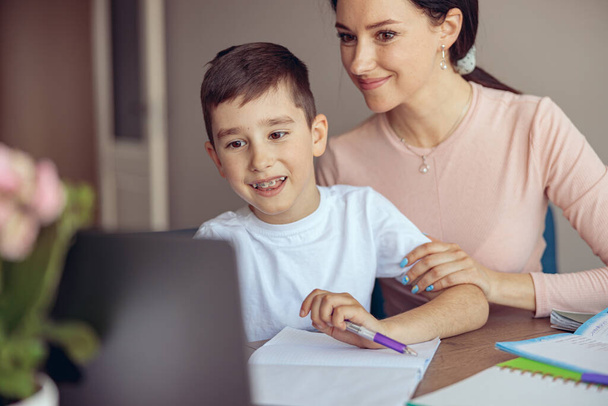 Молодой милый мальчик с брекетами, изучающий домашнее задание на ноутбуке, и мама, помогающая. - Фото, изображение