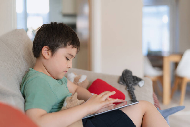 Junge mit Tablet-Spielen im Internet, Kind sitzt auf Sofa und schaut zu oder unterhält sich mit Freund online, Kind entspannt sich morgens im Wohnzimmer, Kinder mit neuem Technologiekonzept - Foto, Bild