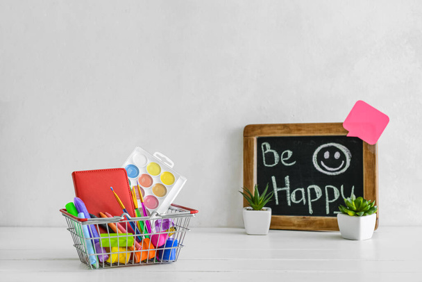 Корзина с различными канцелярскими принадлежностями и мелом доска с фразой быть счастливым на столе возле светлой стены - Фото, изображение
