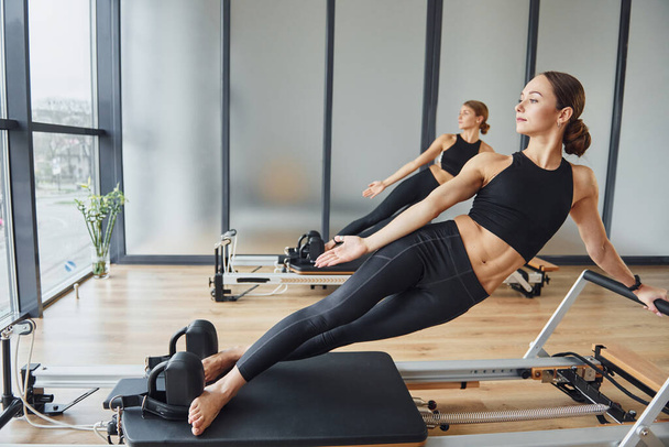 Standing on gym equipment and doing stretches. Two women in sportive wear and with slim bodies have fitness yoga day indoors together. - Photo, image