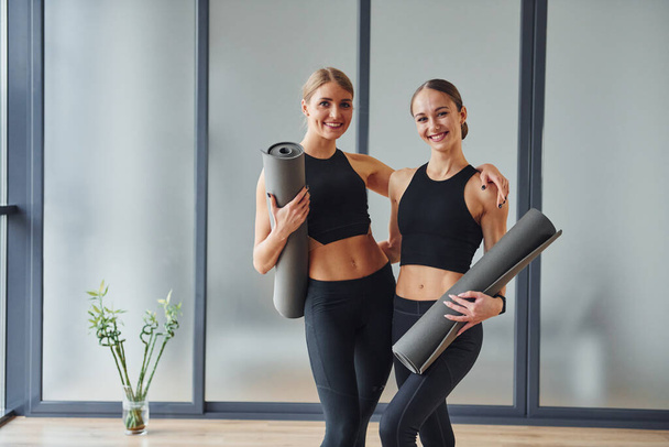 Standing with mats in hands. Two women in sportive wear and with slim bodies have fitness yoga day indoors together. - Foto, Imagen