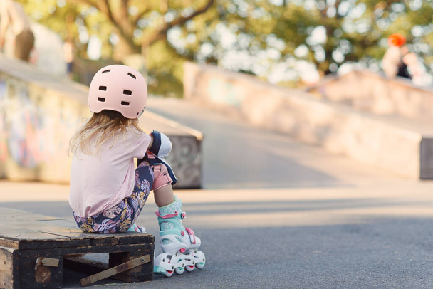 Kleines schönes Mädchen auf Rollschuhen mit Helm und Schutz von Händen und Füßen in einem Skatepark. Fröhliches Vorschulmädchen läuft Schlittschuh an einem sonnigen Tag. - Foto, Bild