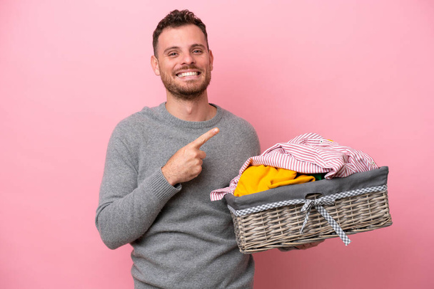 Young Brazilian man holding a clothes basket isolated on pink background pointing to the side to present a product - Foto, Bild