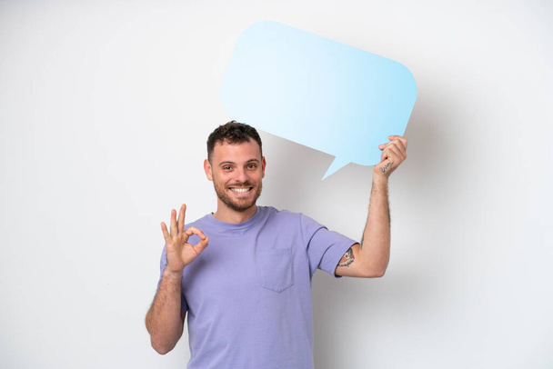 Young Brazilian man isolated on white background holding an empty speech bubble and doing OK sign - Foto, Imagen