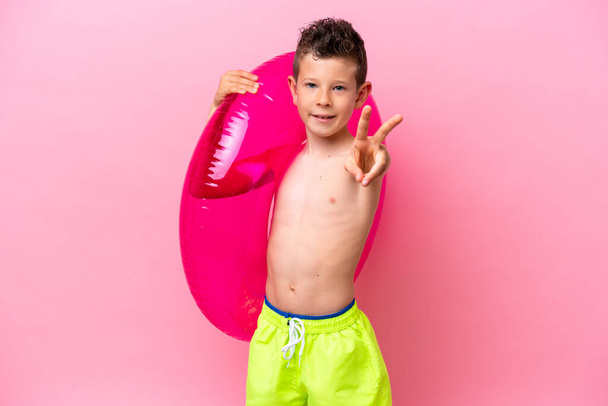 Маленький білий хлопчик тримає надувний пончик ізольований на рожевому тлі, посміхаючись і показуючи знак перемоги
 - Фото, зображення