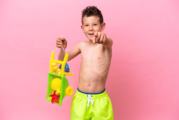 Маленький белый мальчик держит пляжные игрушки изолированы на розовом фоне указывает пальцем на вас с уверенным выражением - Фото, изображение