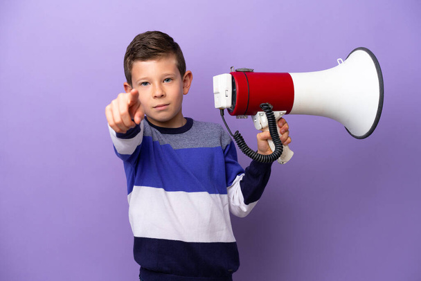 Little boy isolated on purple background holding a megaphone and smiling while pointing to the front - 写真・画像
