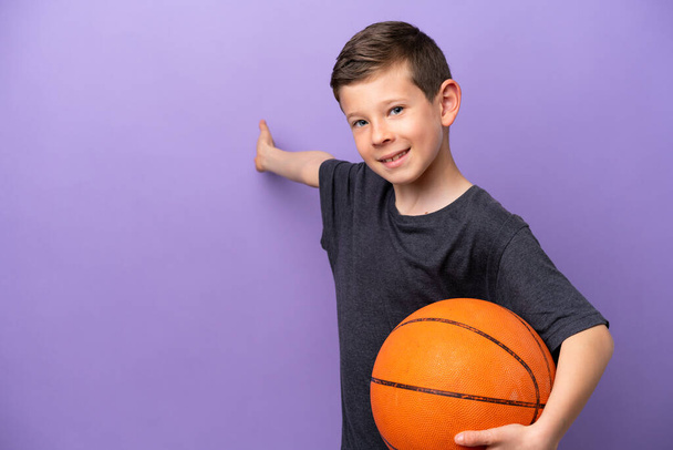 Little boy playing basketball isolated on purple background extending hands to the side for inviting to come - Photo, image