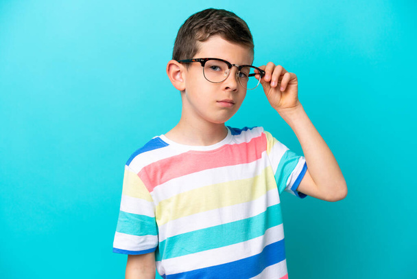 Niño aislado sobre fondo azul con gafas y expresión frustrada - Foto, Imagen