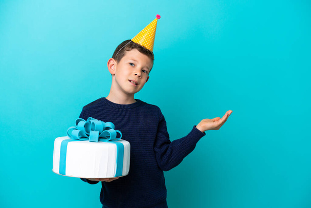 Little boy holding birthday cake isolated on blue background extending hands to the side for inviting to come - Photo, Image