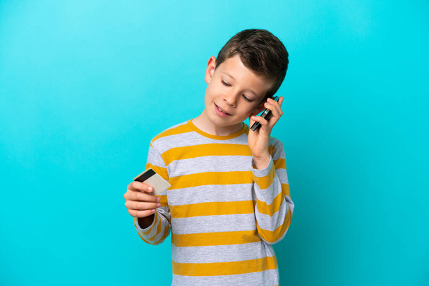 Little boy isolated on blue background keeping a conversation with the mobile phone and holding a credit card - Foto, Bild