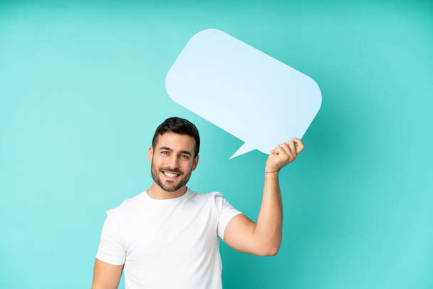 Young handsome caucasian man isolated on blue background holding an empty speech bubble - Foto, afbeelding