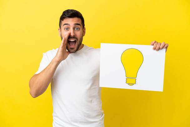 Young handsome caucasian man isolated on yellow background holding a placard with bulb icon and shouting - Zdjęcie, obraz