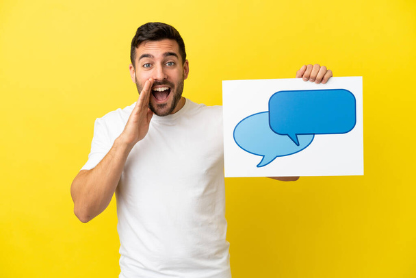 Young handsome caucasian man isolated on yellow background holding a placard with speech bubble icon and shouting - Photo, Image