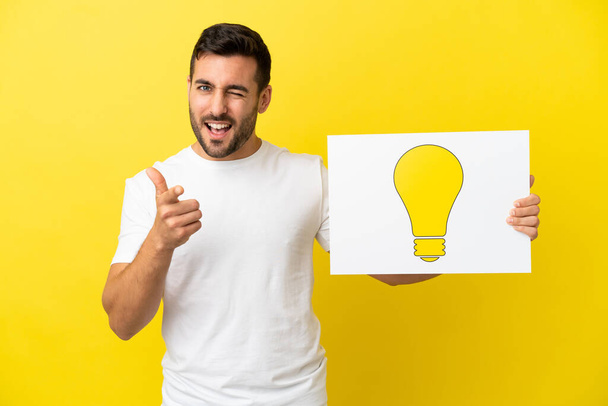 Young handsome caucasian man isolated on yellow background holding a placard with bulb icon and pointing to the front - Foto, Imagem