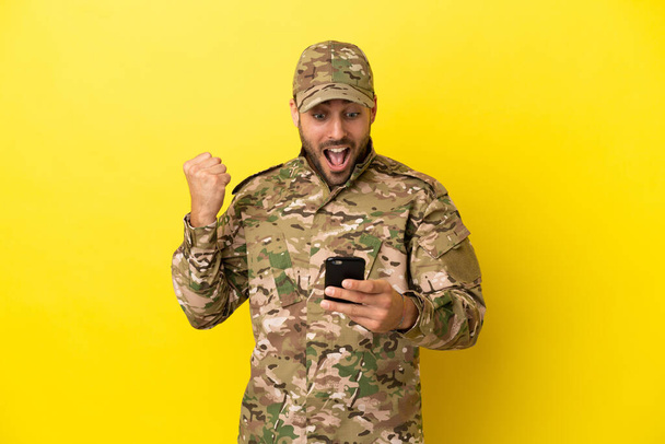 Military man isolated on yellow background surprised and sending a message - Φωτογραφία, εικόνα