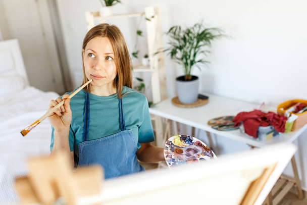 Woman artist holding paintbrush.Creative woman enjoying painting.Young beautiful woman in art studio, taking colorful paints from palette,creating masterpiece on easel, work, imagination. - Photo, Image