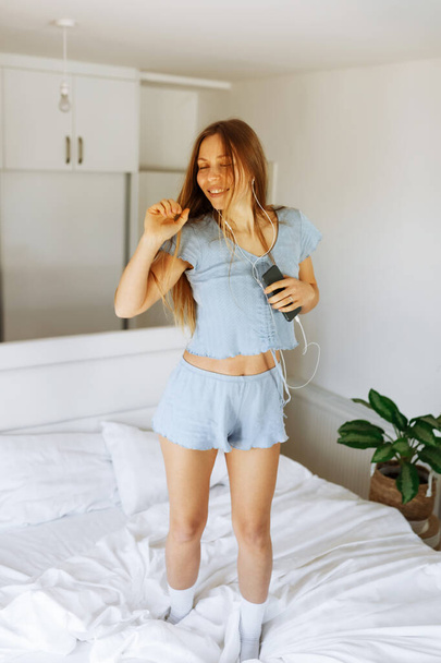 Young teen girl in pajama standing on her bed and singing a song. Cheerful caucasian adolescent girl with headphones dancing on her bedroom  - Foto, immagini