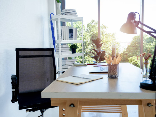 Moderne werkplaats met lege zwarte stoel, potlood, notebook tafellamp en laptop computer op houten bureau in de buurt van het boekenrek en glazen raam, kantoor, wit . - Foto, afbeelding