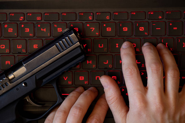 A gun lies on a black laptop keyboard with red backlight and a woman's hand on the keyboard, security threat, cyber attack, system hacking, work online - Foto, Imagem