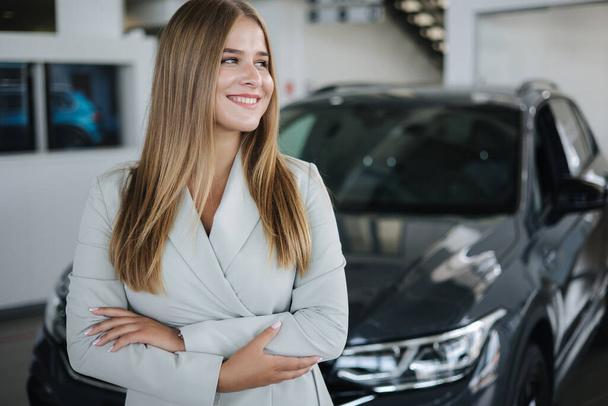 Портрет продавца в автосалоне. Привлекательная молодая женщина стоит перед машиной. Красивая женщина в костюме перед роскошной машиной - Фото, изображение