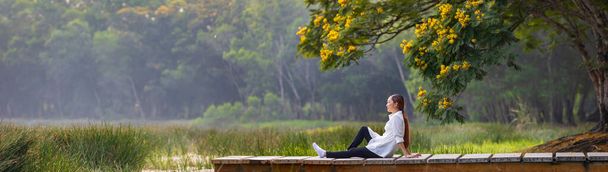 Panorama of asian woman sitting on the edge of dock with peaceful natural park during summer with yellow flower blossom for serene and relaxation outdoor recreation - Fotó, kép