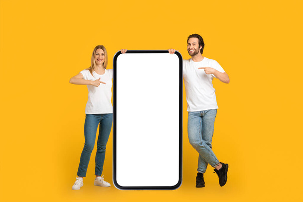 Pareja europea milenaria satisfecha en camisetas blancas señala con el dedo el enorme teléfono con la pantalla vacía - Foto, imagen