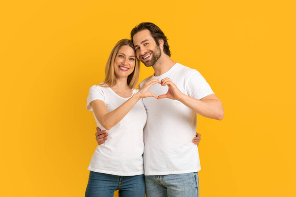 Happy attractive millennial european wife and husband with stubble in white t-shirts make heart with hands - Foto, Imagem