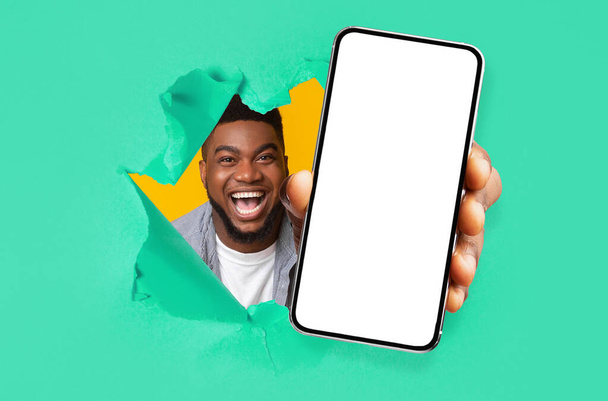 Ικανοποιημένος ενθουσιασμένοι χιλιετή αφροαμερικανός άνθρωπος ματιά μέσα από την τρύπα στο πράσινο χαρτί και δείχνει smartphone - Φωτογραφία, εικόνα