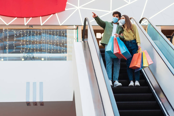 Ζευγάρι ψώνια φορώντας μάσκες στέκεται σε κινούμενες σκάλες στο εμπορικό κέντρο - Φωτογραφία, εικόνα