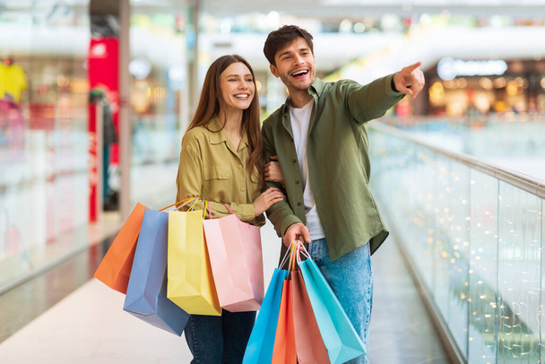 Cónyuges felices sosteniendo bolsas de comprador señalando las compras de dedo en el hipermercado - Foto, imagen