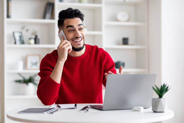 Froh junger arabischer Mann mit Bart telefoniert mit Klient am Arbeitsplatz mit Laptop im Home Office Interieur - Foto, Bild