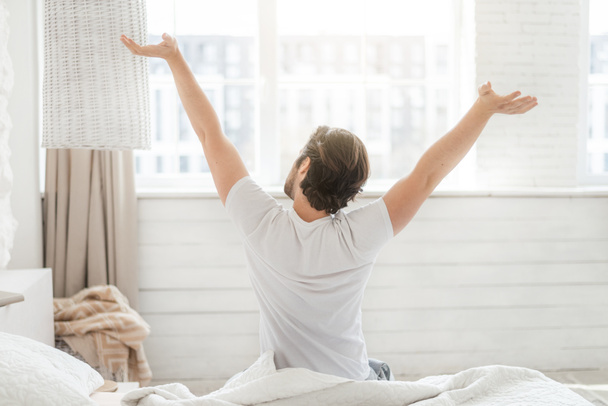 Happy millennial caucasian guy wake up and stretching body, sitting on soft bed on window background - Fotó, kép