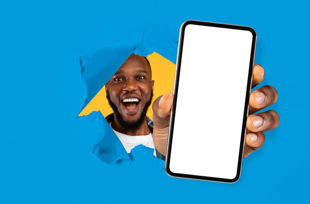 Ευτυχισμένο ενθουσιασμένοι χιλιετή αφροαμερικανός άνδρας με ανοιχτό στόμα κοιτάζει μέσα από την τρύπα στο μπλε χαρτί και δείχνει smartphone - Φωτογραφία, εικόνα