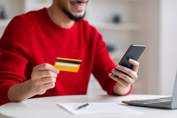 Smiling young arab guy with beard showing credit card and typing on smartphone in home office interior - Photo, Image