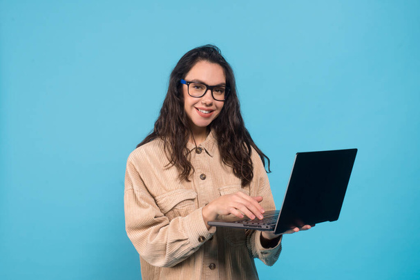 Smiling millennial european woman brunette student in glasses typing on laptop isolated on blue background - Foto, immagini