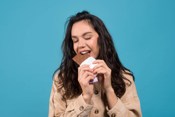 Happy young european brunette woman with closed eyes eats chocolate and enjoys taste of sweets - Valokuva, kuva