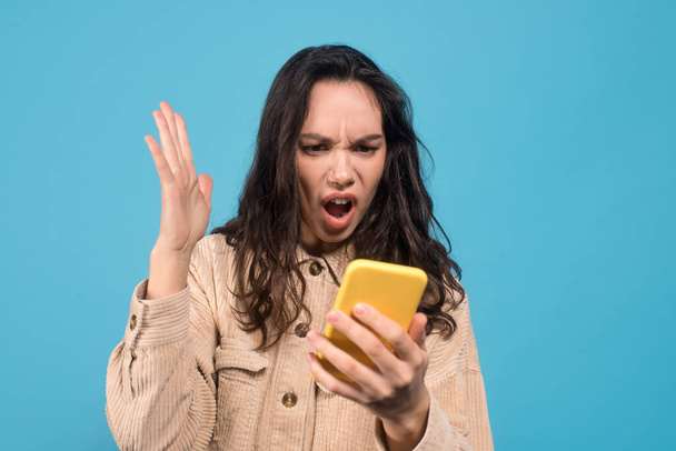 Sad shocked young european brunette woman with open mouth looks at smartphone isolated on blue background - Photo, Image
