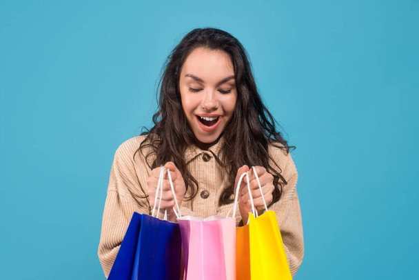 Surprised glad young caucasian brunette lady looking into bags with purchases, isolated on blue background - Photo, image