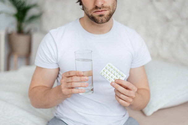 Unhappy millennial caucasian guy with stubble wake up, sits on bed, holds glass of water and pills in bedroom - Foto, imagen