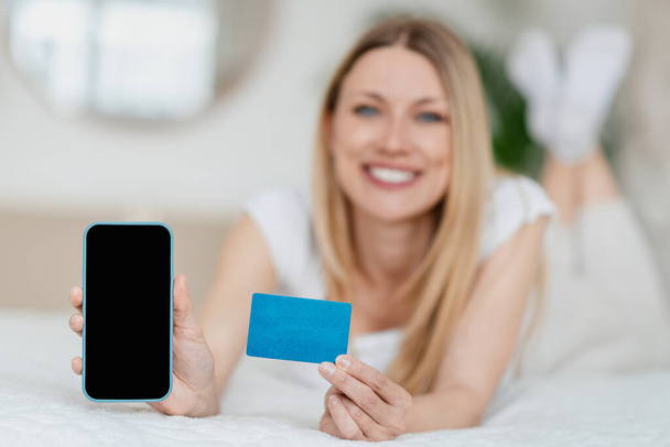 Счастливая молодая блондинка европейская леди показывает смартфон с чистым экраном и кредитной картой, лежит на кровати в спальне - Фото, изображение