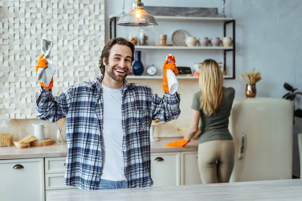 Jovem mulher europeia feliz e marido em luvas de borracha levantar as mãos com suprimentos de limpeza, poeira limpa na cozinha - Foto, Imagem